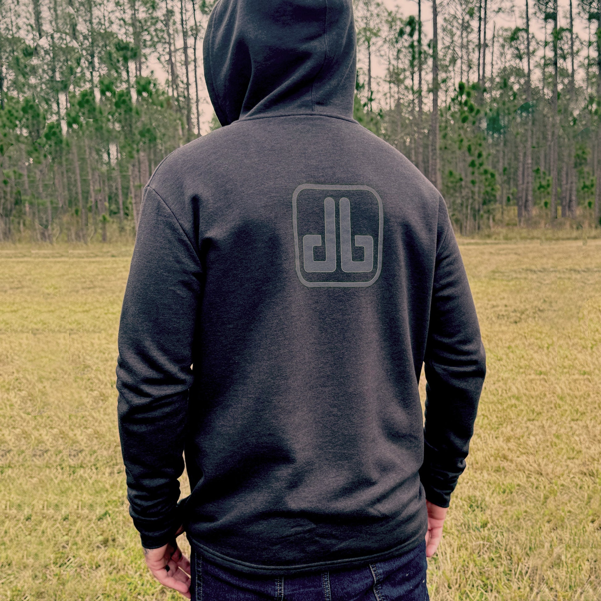 Person wearing a dark hoodie with a db ogo on the back, standing in an open field with trees in the background.